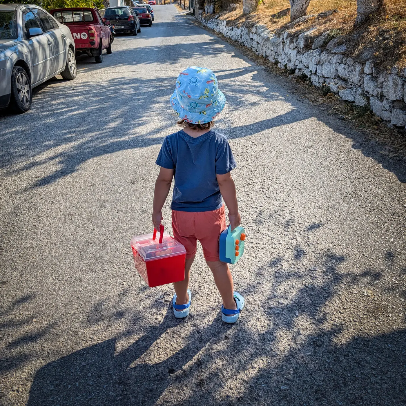 A toddler in a sun hat walking down a village road, carrying a Koobiba