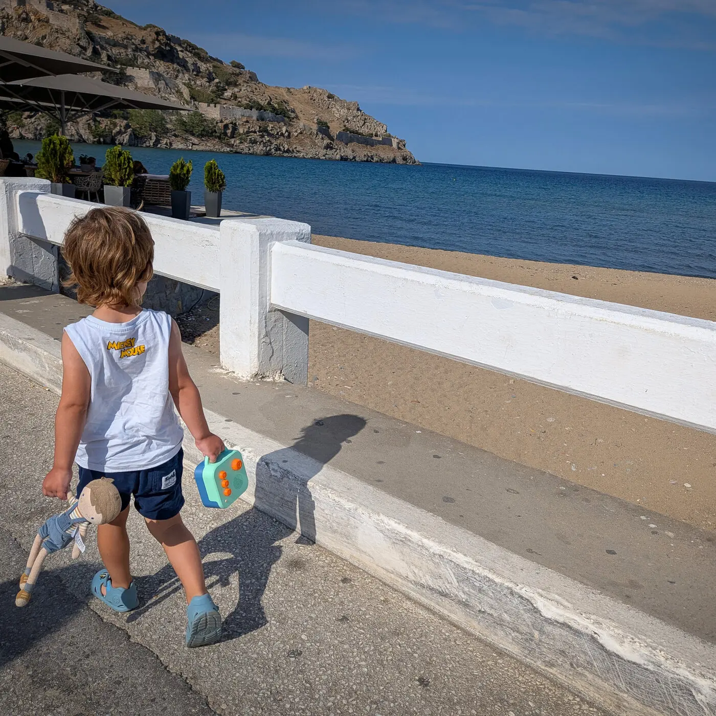 A toddler walking along a seafront path, carrying a Koobiba