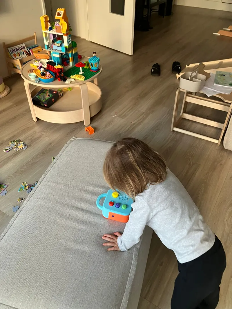 Child leaning on a couch, holding a blue Koobiba audio player by its handle